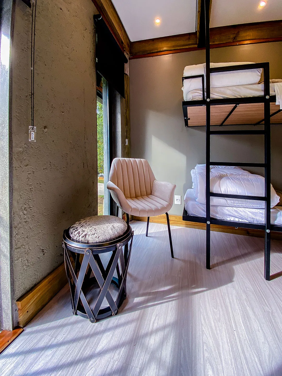 Canto do quarto com poltrona rosada, banqueta de metal e beliche com colchões e roupa de cama brancos.