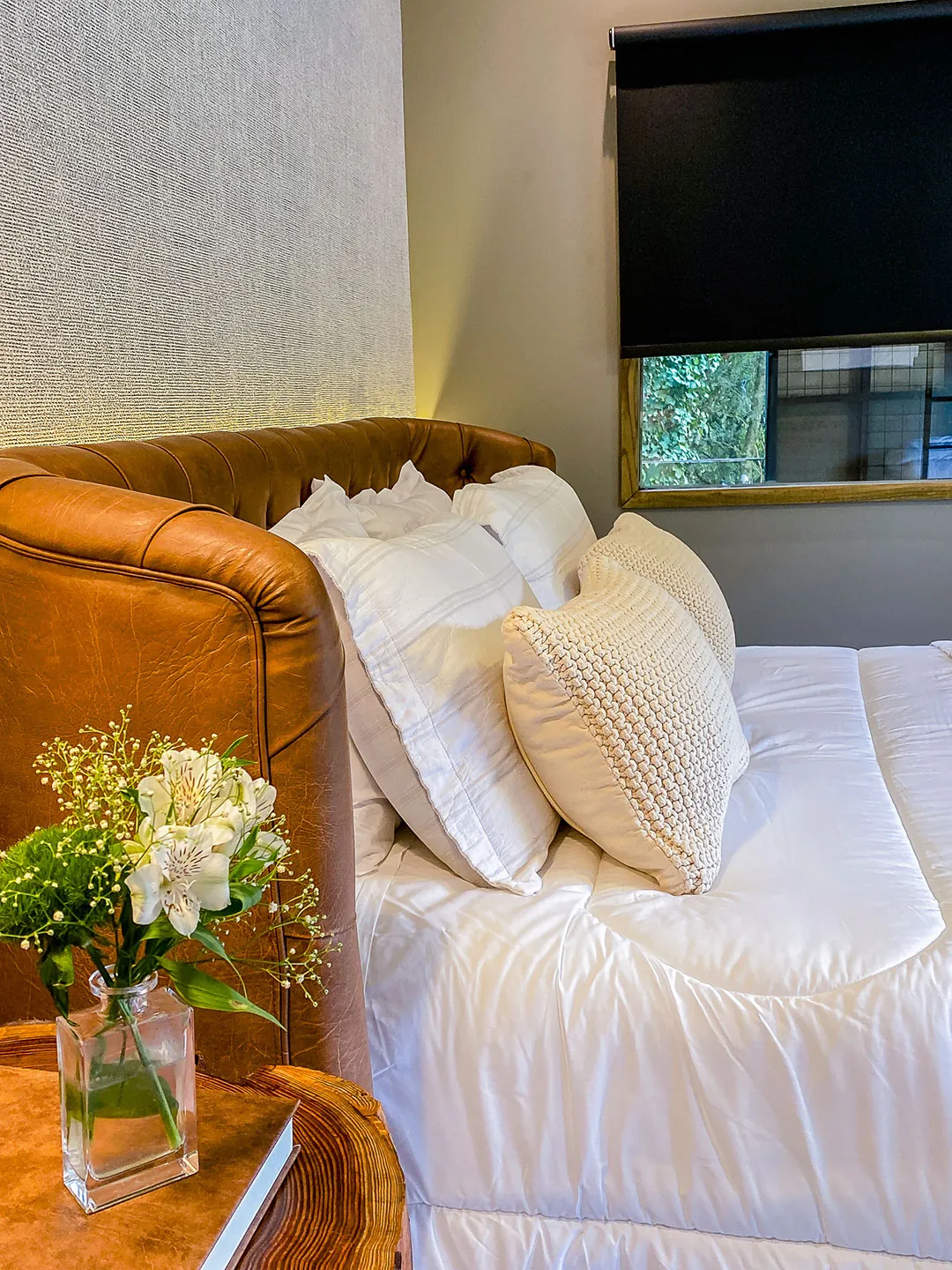 Detalhe da cama com cabeceira de couro caramelo, travesseiros brancos, almofada de tricô e vaso de flores sobre mesa de tronco.