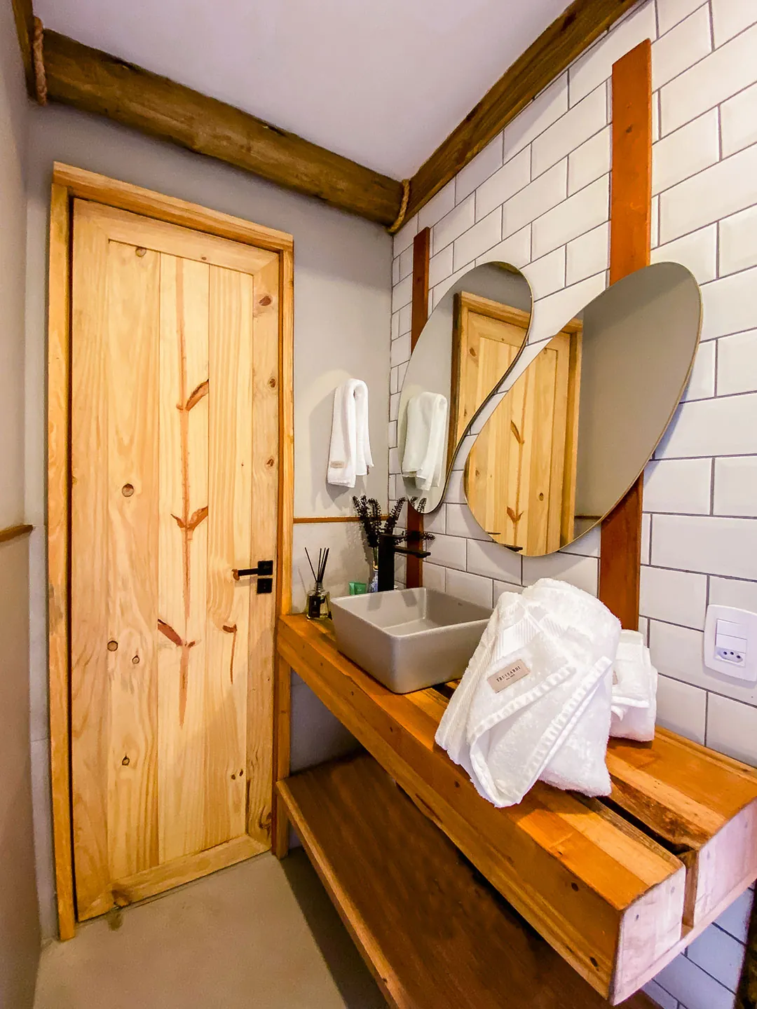 Banheiro com porta de madeira clara, bancada de madeira com cuba branca, roupão dobrado, espelhos ovais e parede de azulejos brancos.