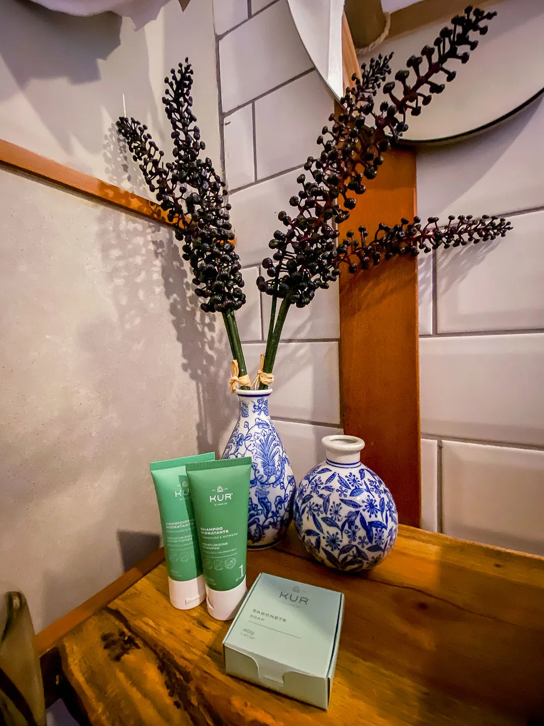 Detalhe da bancada de madeira com dois vasos azuis e brancos, galhos secos, bisnagas e caixa de amenities verdes.