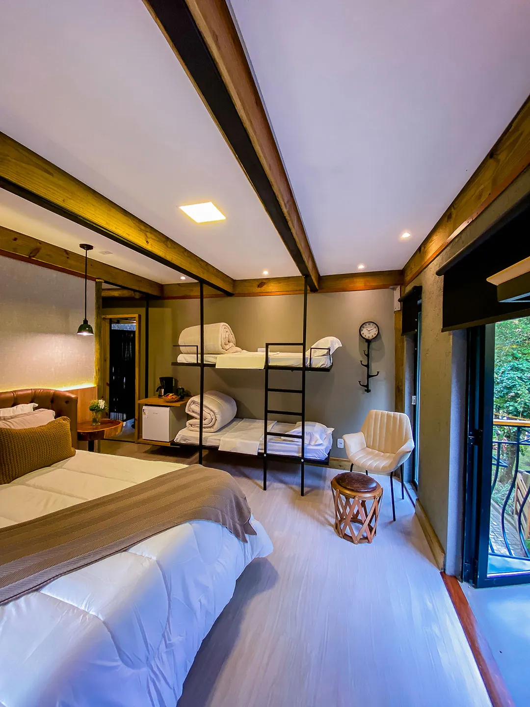 Vista ampla do quarto a partir da cama: beliche de metal, poltrona, banqueta, frigobar e porta de vidro para a sacada; teto branco com vigas de madeira.