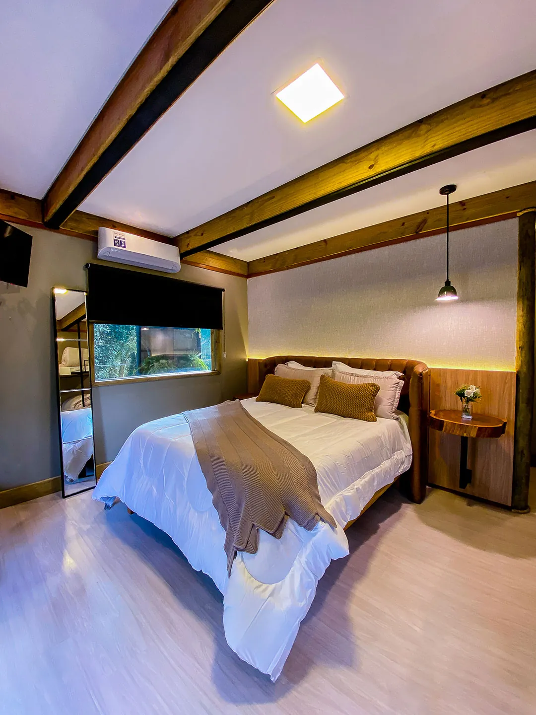 Cama de casal com manta marrom e almofadas caramelo, cabeceira estofada, mesa lateral de madeira, luminária pendente, janela e ar-condicionado.