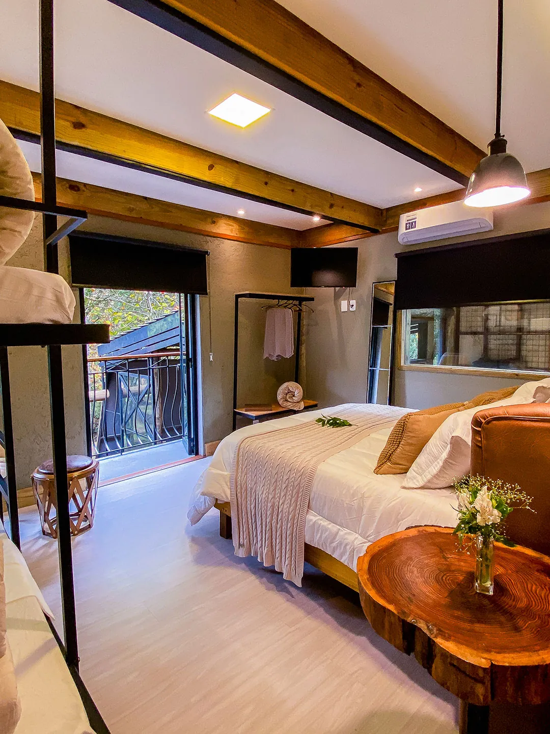Cama de casal vista de lado com mesa de tronco e flores, beliche à esquerda, arara com roupão e porta aberta para a sacada com gradil.