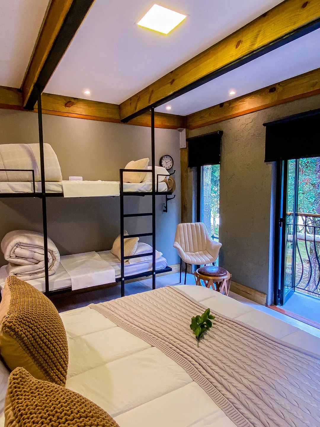 Beliche de metal com colchões, cobertores e travesseiros brancos, poltrona e porta de vidro para sacada; cama com manta e almofadas de tricô em primeiro plano.