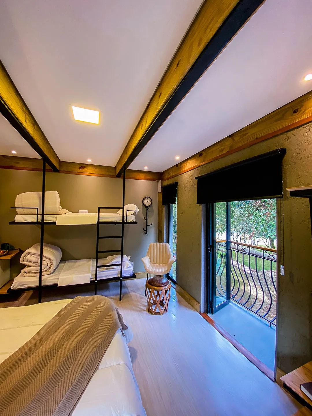 Beliche de metal com cobertores dobrados, poltrona, banqueta e porta de vidro aberta para a sacada; canto da cama em primeiro plano.