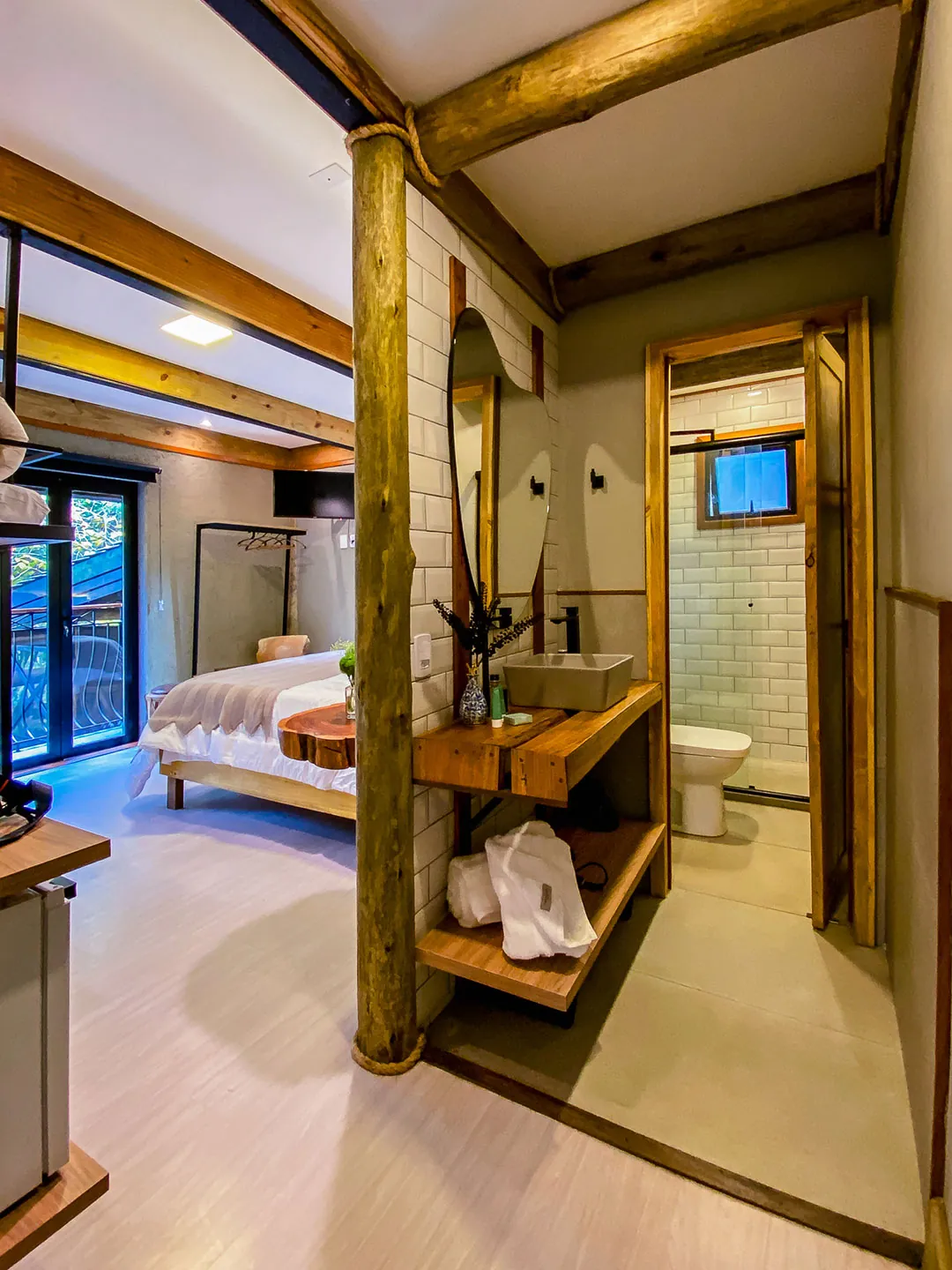 Coluna de tronco e bancada de madeira com cuba e roupões; ao fundo, porta aberta com vaso sanitário e parede de azulejos; cama visível à esquerda.