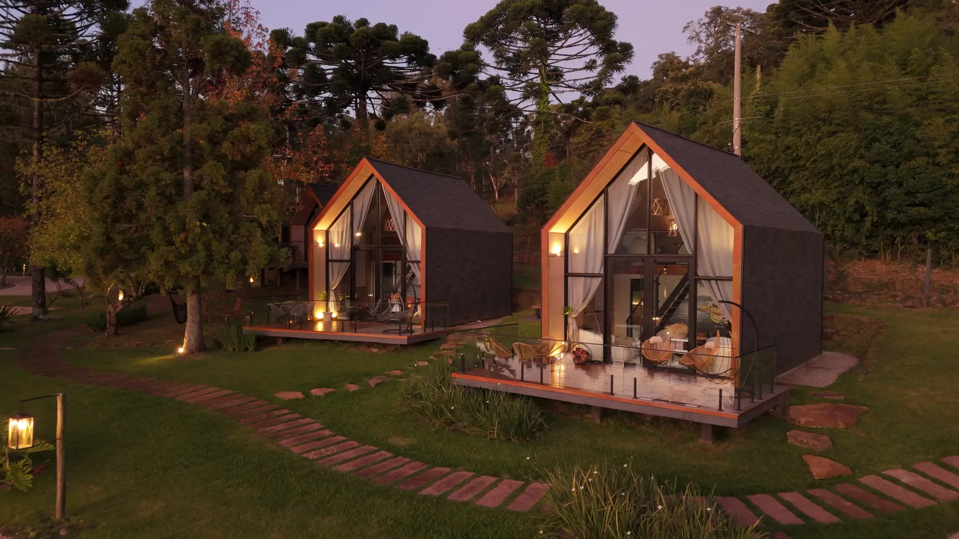 Duas lake houses com fachada de vidro e luzes acesas ao anoitecer, com araucárias ao fundo